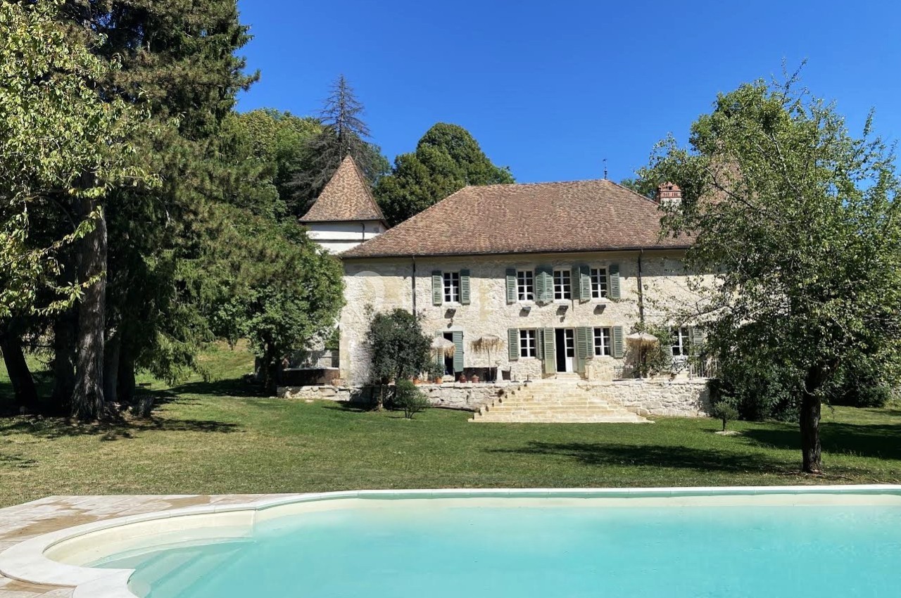 Maison en pierre avec toit en tuiles, entourée d'arbres, avec une piscine au premier plan sous un ciel bleu.