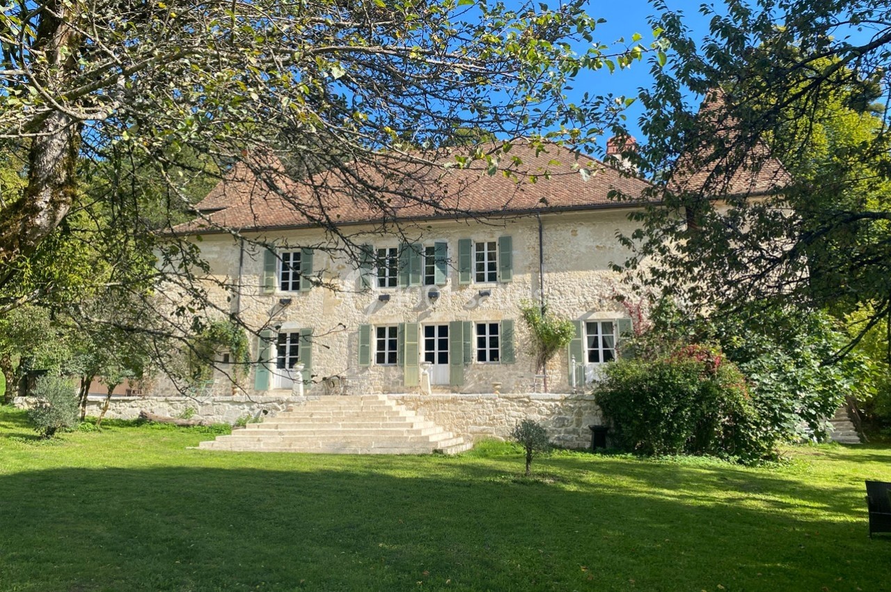 Grande maison en pierre avec volets verts, entourée d'arbres et d'une pelouse, sous un ciel bleu clair.