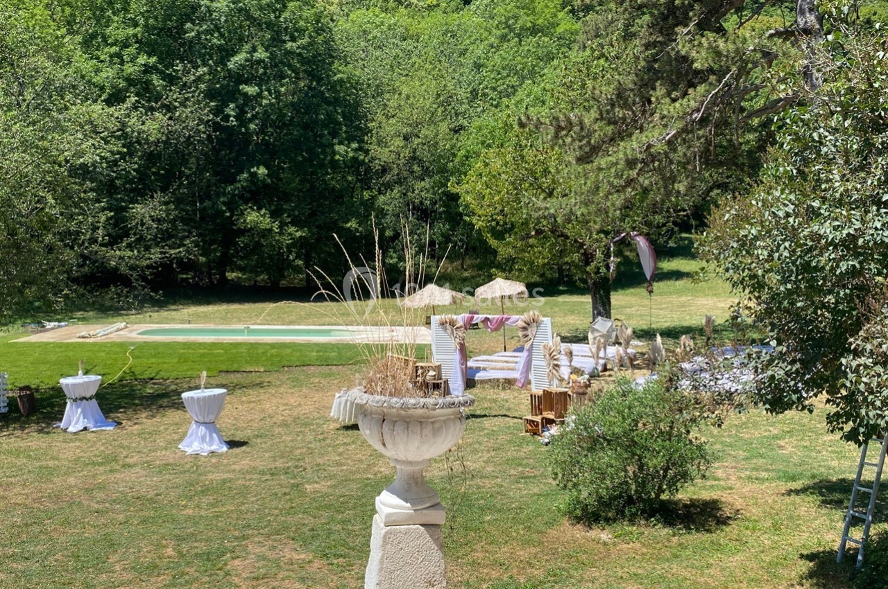 Jardin avec piscine, tables dressées et chaises en bois, entouré de verdure et d'arbres sous un ciel clair.