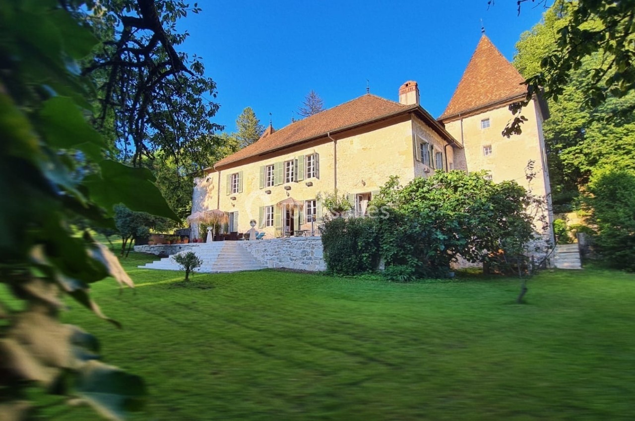 Grande maison en pierre avec tour, entourée d'un jardin verdoyant sous un ciel bleu clair.