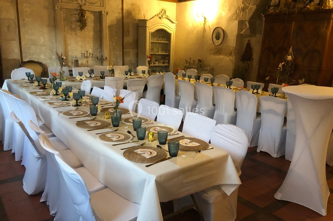 Salle décorée pour un repas, avec longues tables dressées, nappes blanches, vaisselle élégante et chaises houssées.