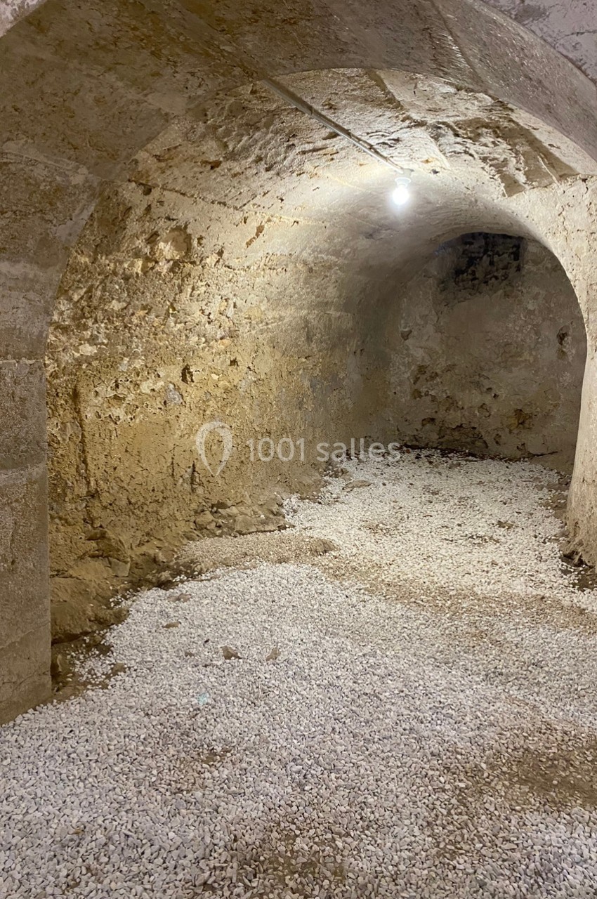 Cave voûtée en pierre avec sol recouvert de gravier, éclairée par une ampoule suspendue.