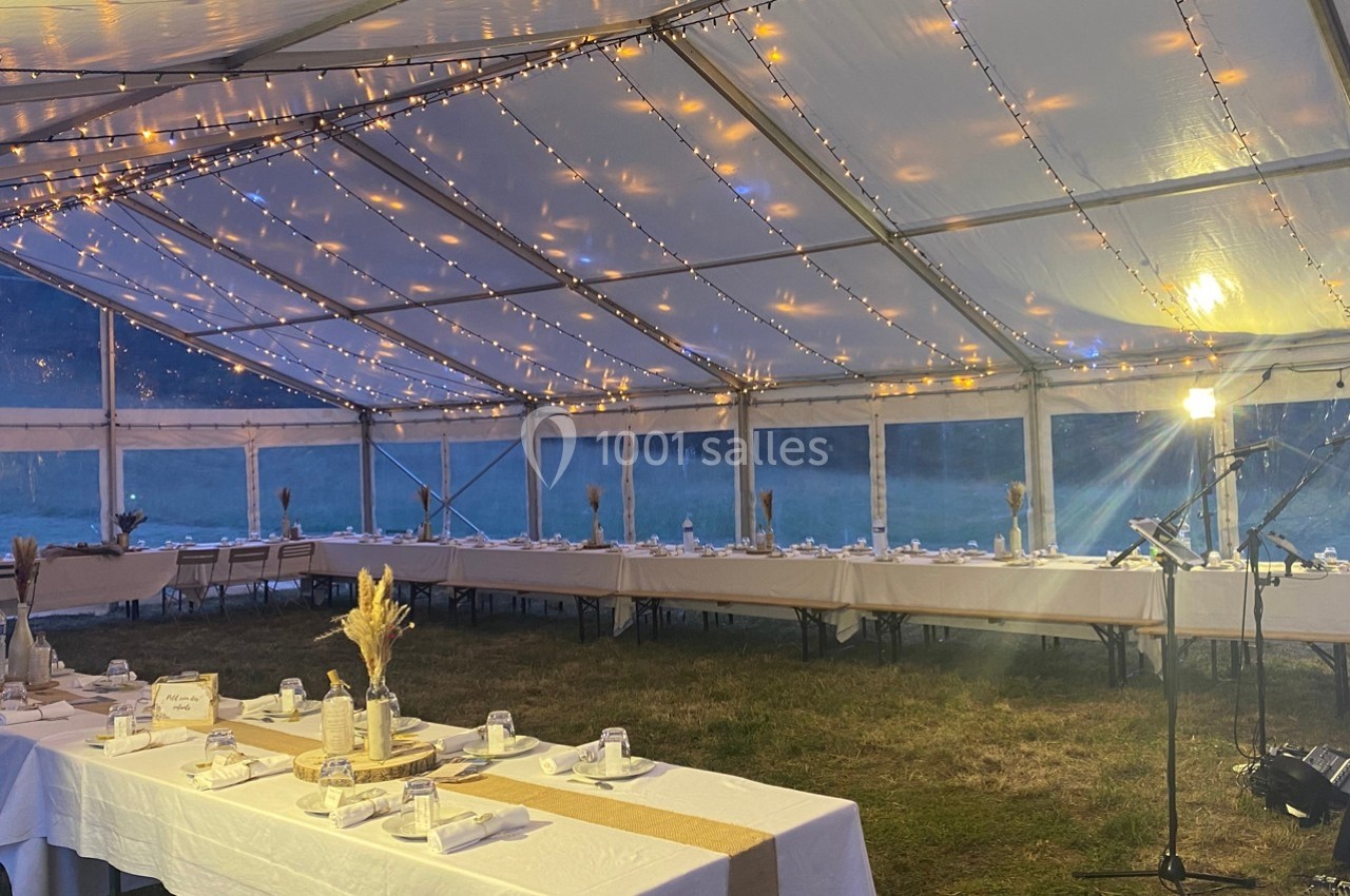 Grande tente éclairée par des guirlandes lumineuses, aménagée avec des tables décorées pour un événement en soirée.