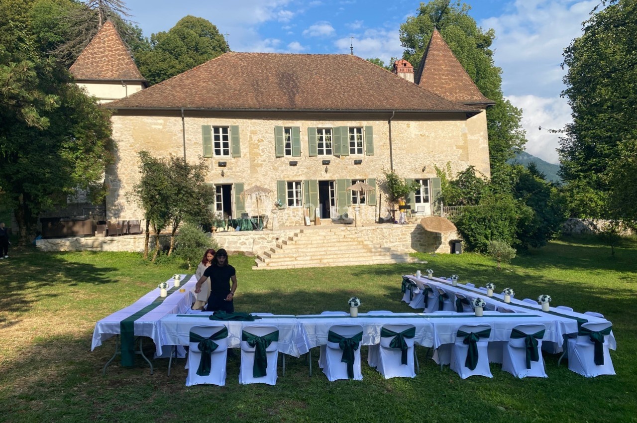 Tables dressées en extérieur devant une grande maison en pierre entourée de verdure, par une journée ensoleillée.