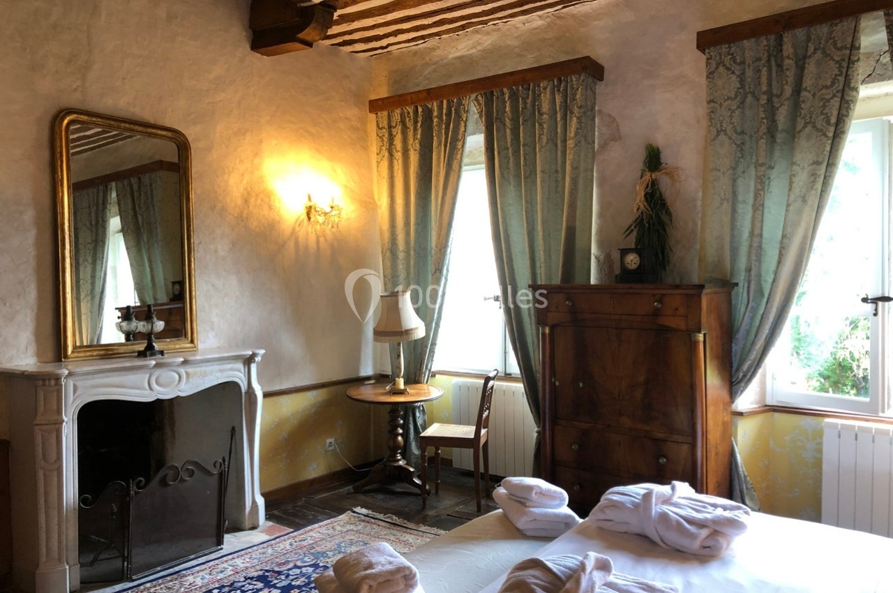 Chambre lumineuse avec lit, cheminée, armoire en bois, miroir, rideaux élégants et poutres apparentes au plafond.