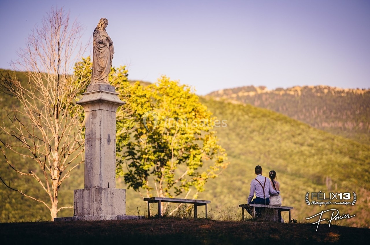 Statue religieuse sur un socle en pierre, avec deux personnes assises sur un banc face à un paysage de montagne.