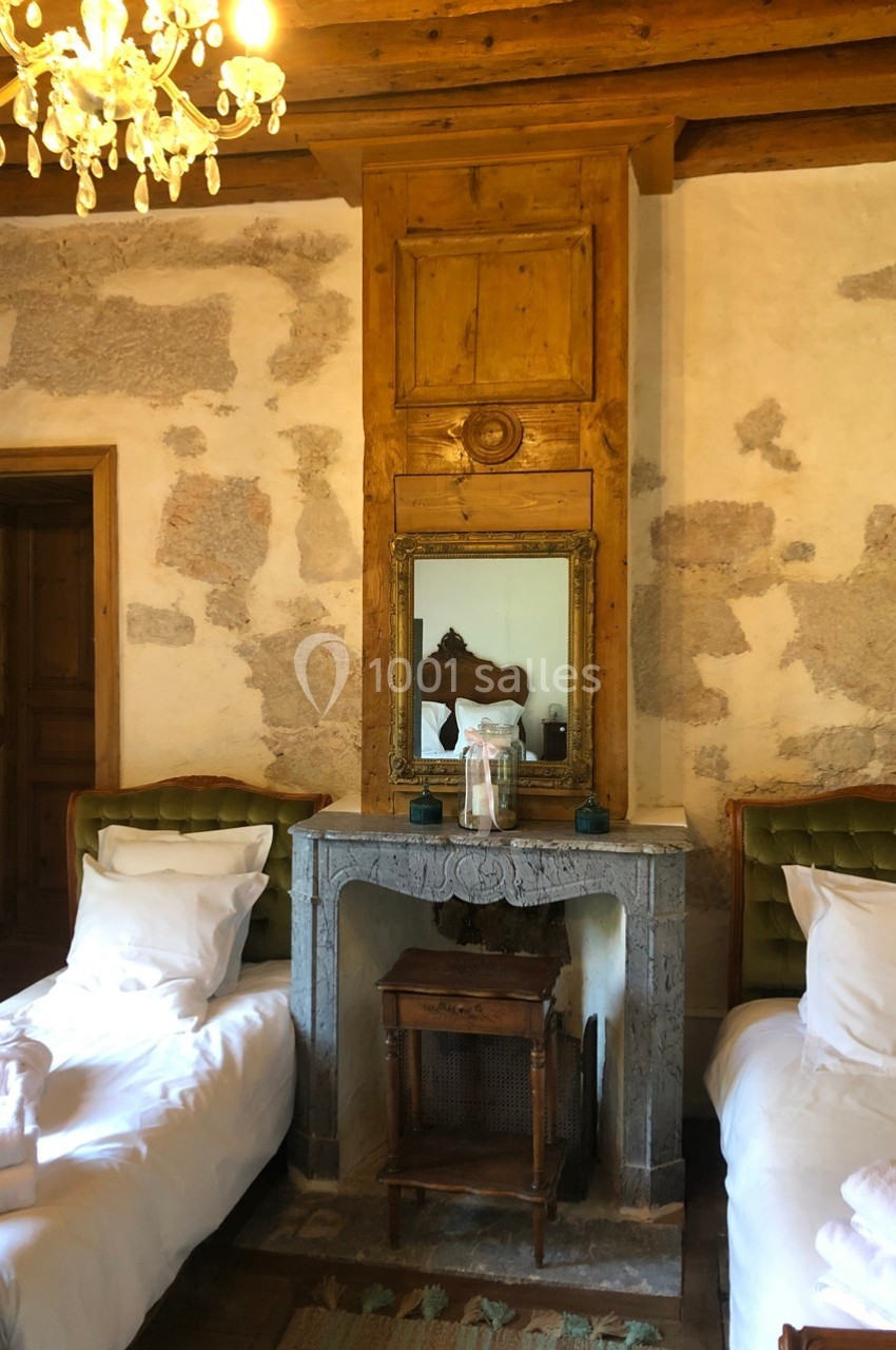 Chambre rustique avec deux lits simples, cheminée en pierre, miroir encadré de bois et mur en pierres apparentes.