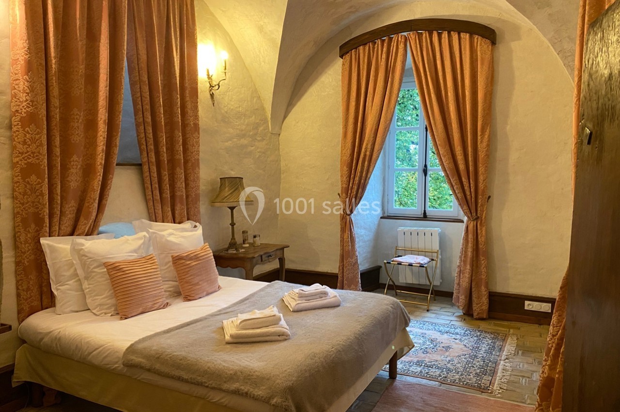 Chambre avec lit double, rideaux orange, murs en pierre, lampe de chevet et fenêtre donnant sur un jardin verdoyant.