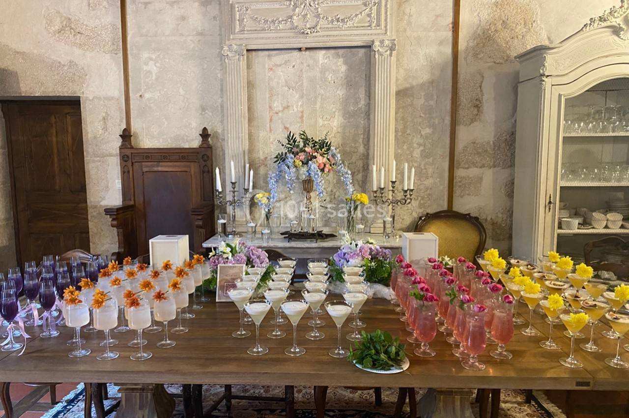 Table en bois garnie de cocktails colorés, décorée de fleurs et entourée de mobilier ancien dans une salle élégante.