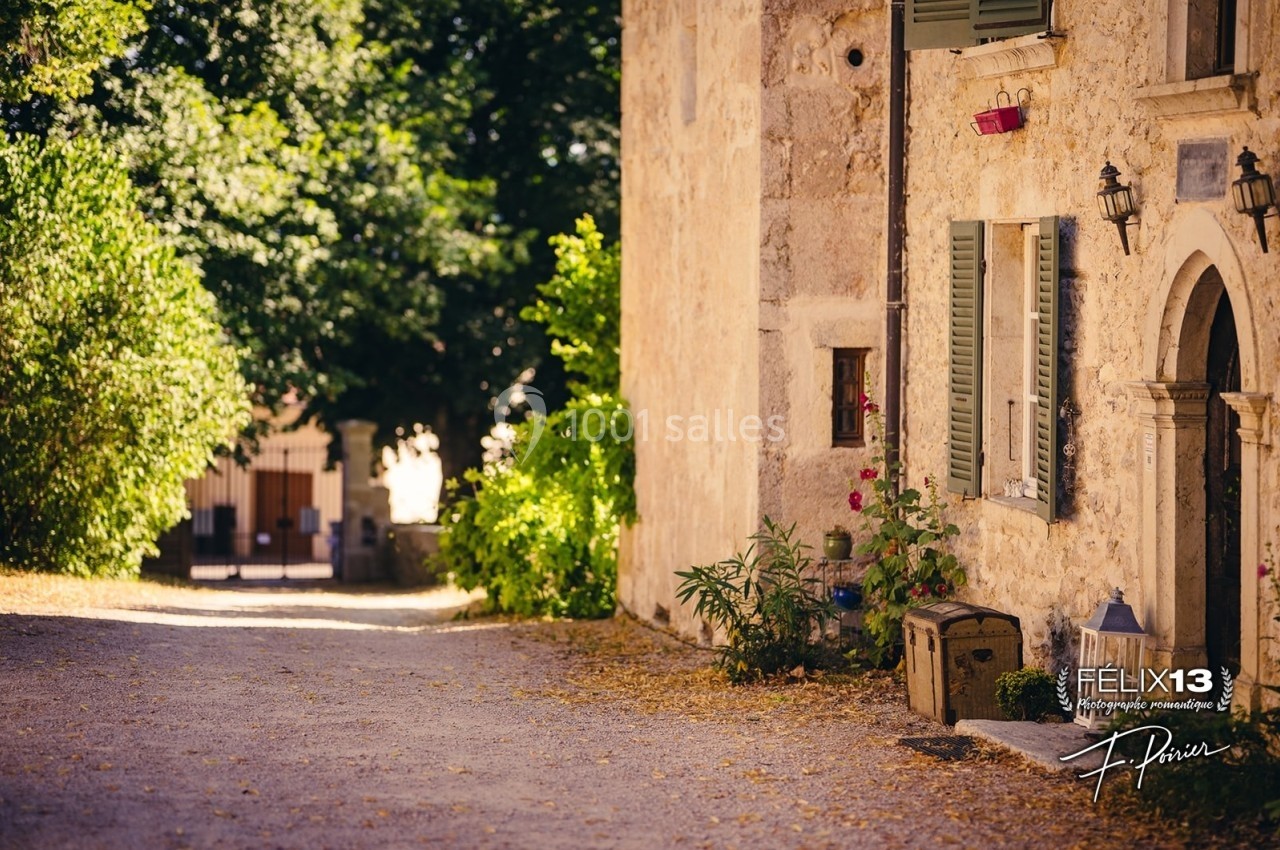 Rue pavée bordée de végétation et d'une maison en pierre avec volets verts, éclairée par une lumière douce.