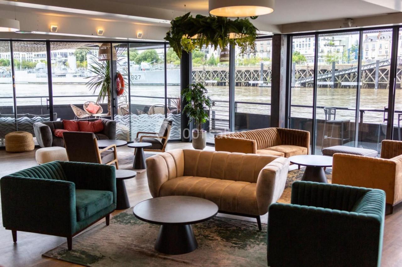 Espace lounge lumineux avec fauteuils, canapés et tables basses, vue sur une rivière à travers de grandes baies vitrées.