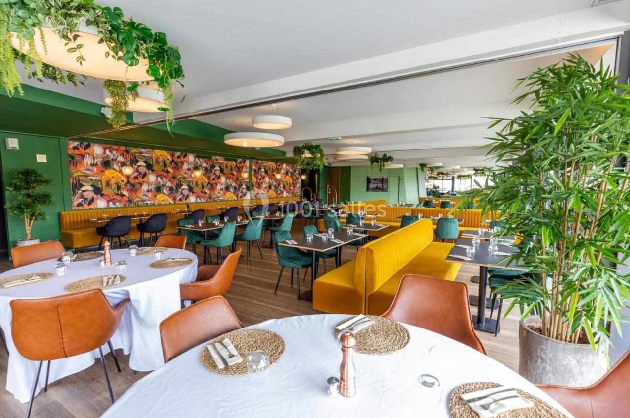 Salle de restaurant lumineuse avec tables dressées, banquettes jaunes, plantes décoratives et fresque colorée au mur.