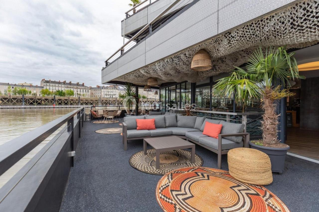 Terrasse aménagée avec canapé, tables et plantes, donnant sur un fleuve et des bâtiments en arrière-plan.