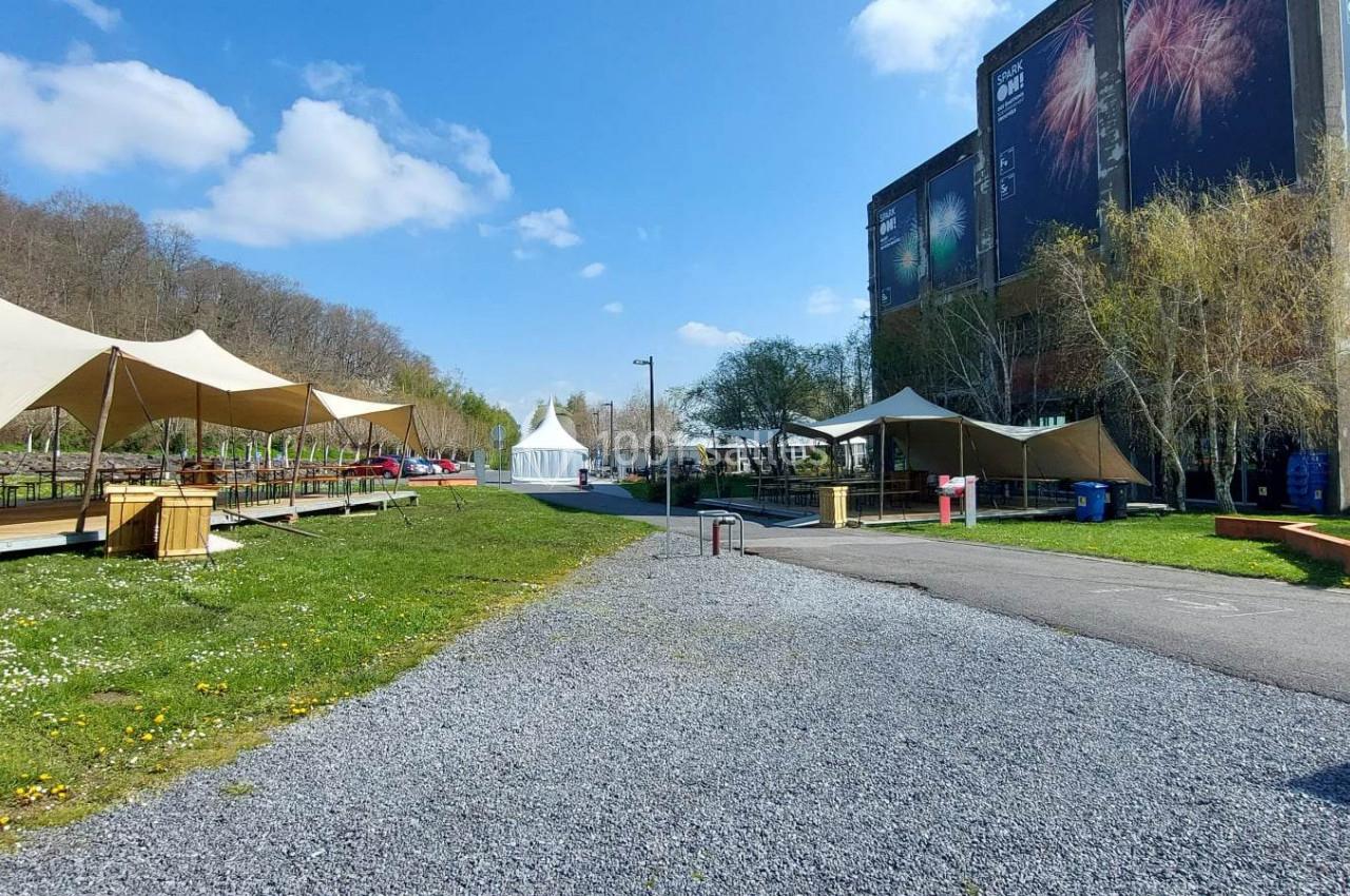 Allée bordée de tentes et d'espaces verts, avec un bâtiment industriel en arrière-plan sous un ciel dégagé.