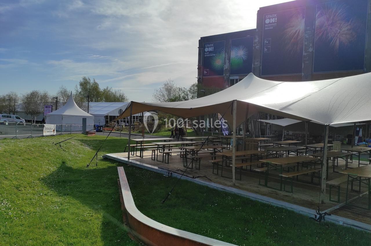 Terrasse extérieure avec tables et bancs sous des tentes, située près d'un bâtiment et entourée d'espaces verts.