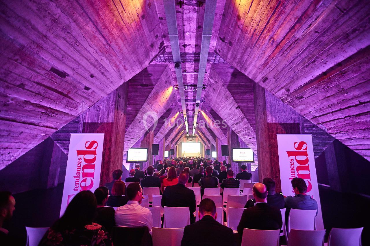Salle de conférence avec un public assis, éclairage violet et écrans diffusant une présentation.