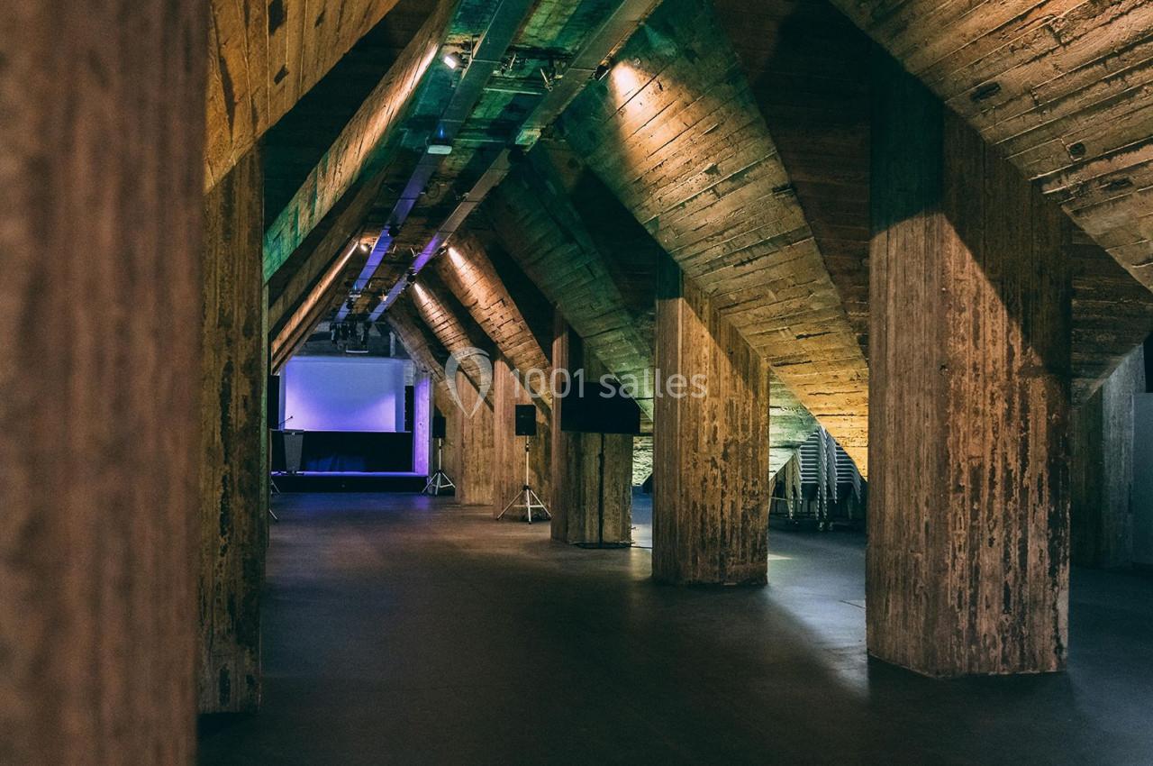 Vue intérieure d'une salle avec des colonnes en bois brut, un éclairage tamisé et une scène visible au fond.