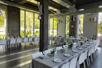 Miniature Mariage Salle lumineuse avec de grandes baies vitrées, tables dressées avec nappes grises, vaisselle et décoration florale.