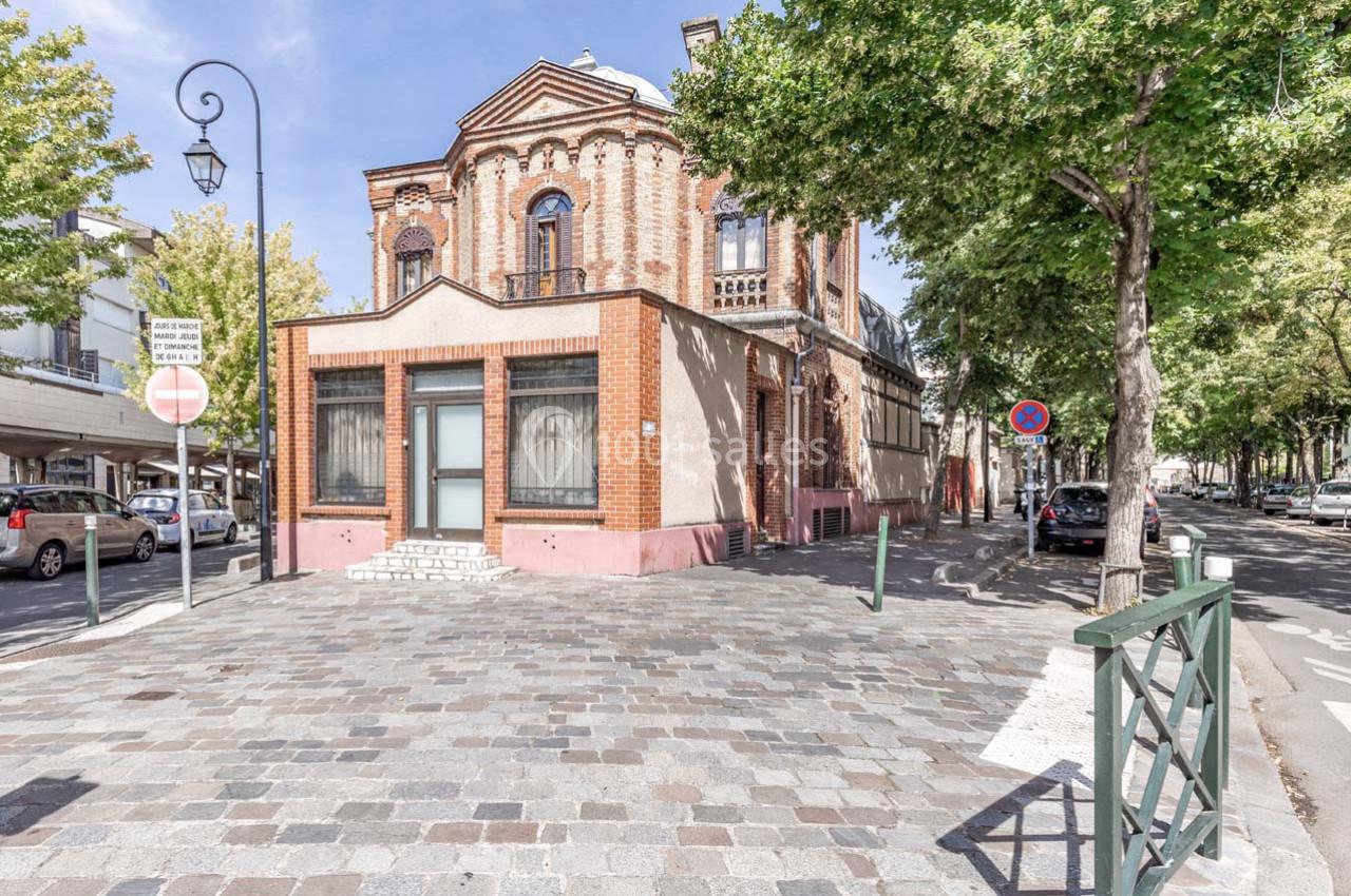 Bâtiment en briques rouges à angle arrondi, bordé d'arbres et situé à l'intersection de rues pavées.