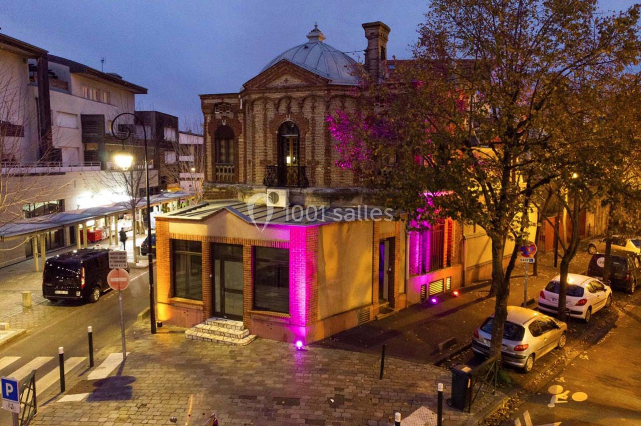 Vue d'un bâtiment en briques illuminé de lumières violettes, entouré de rues pavées et d'arbres en automne.
