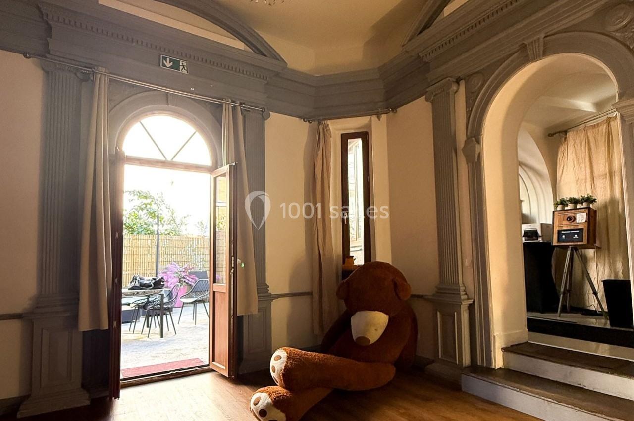 Un grand ours en peluche est posé contre un mur dans une pièce lumineuse avec une porte ouverte sur une terrasse.