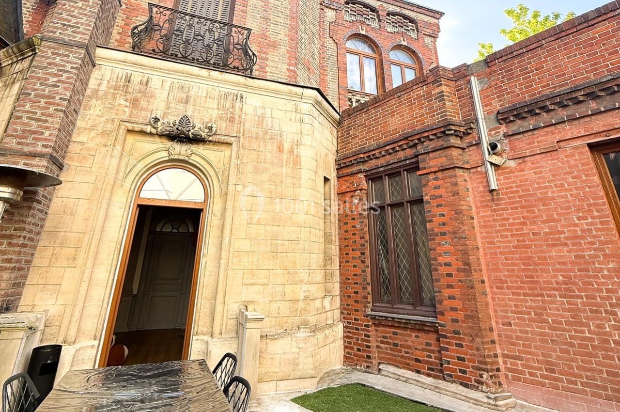 Façade en briques rouges et pierre avec une porte voûtée, fenêtres à croisillons et terrasse aménagée.