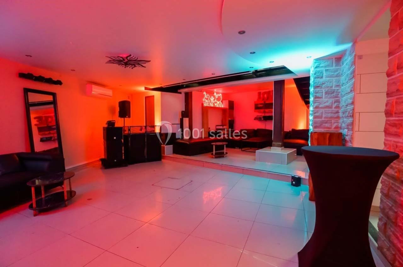 Salle moderne avec éclairage coloré, mobilier contemporain, canapés noirs et murs en pierre partielle.