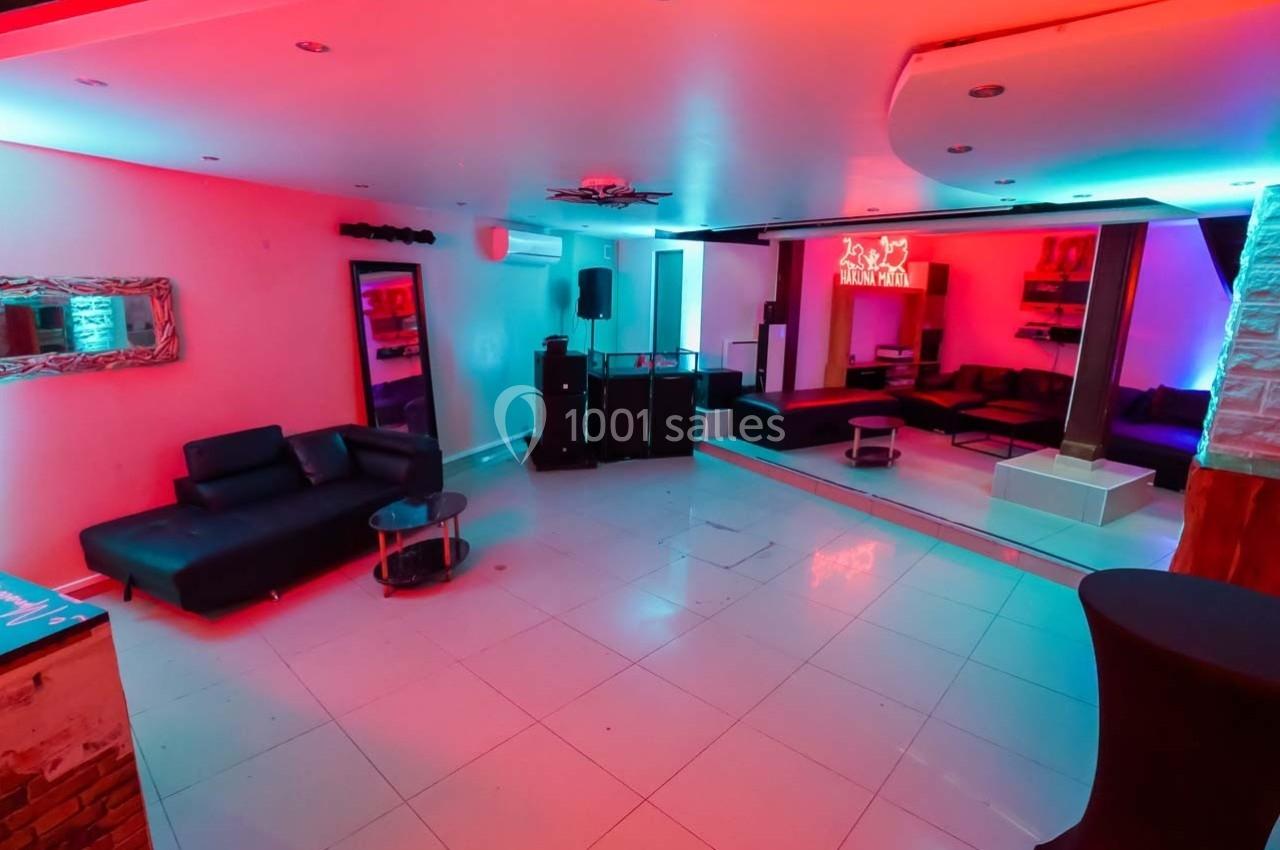 Salle moderne avec éclairage LED coloré, canapés noirs, tables basses et espace ouvert sur sol carrelé blanc.