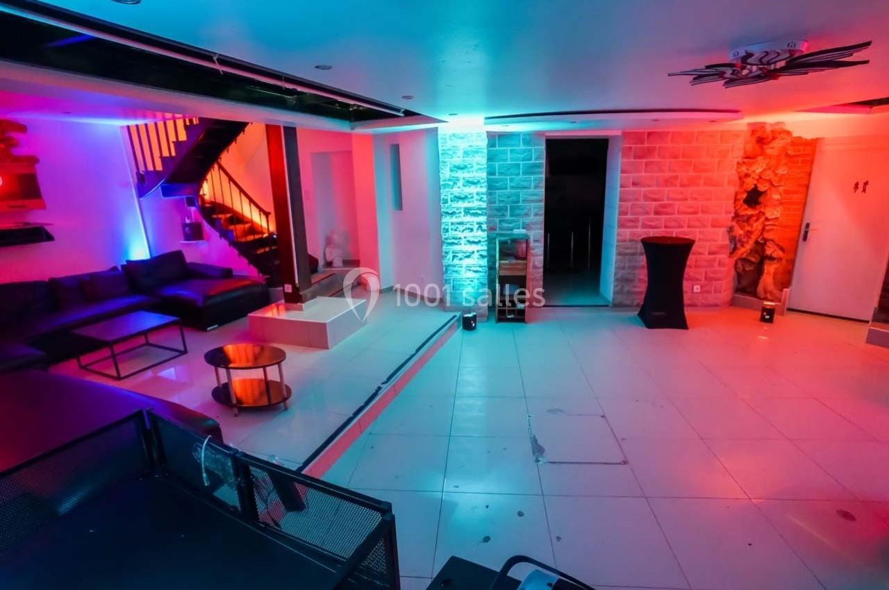 Salle moderne avec éclairage coloré, canapé d'angle, table basse, murs en pierre et escalier visible en arrière-plan.