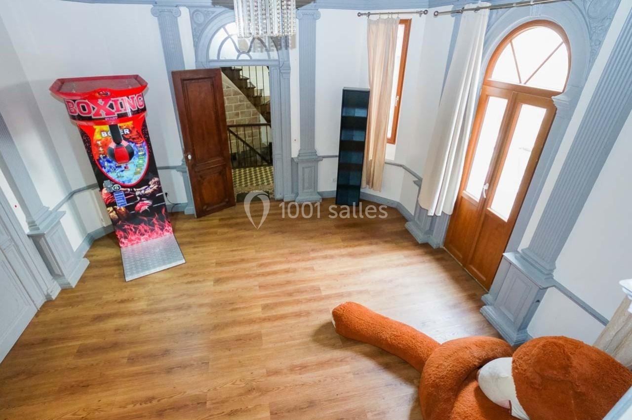 Salle lumineuse avec un sol en bois, une borne de jeu de boxe, un grand fauteuil orange et des portes-fenêtres.