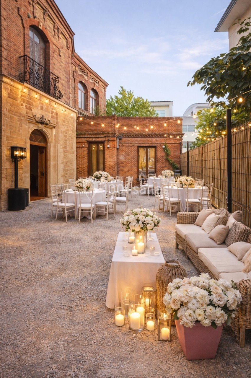 Cour extérieure aménagée pour un événement, avec tables décorées, guirlandes lumineuses et bougies.