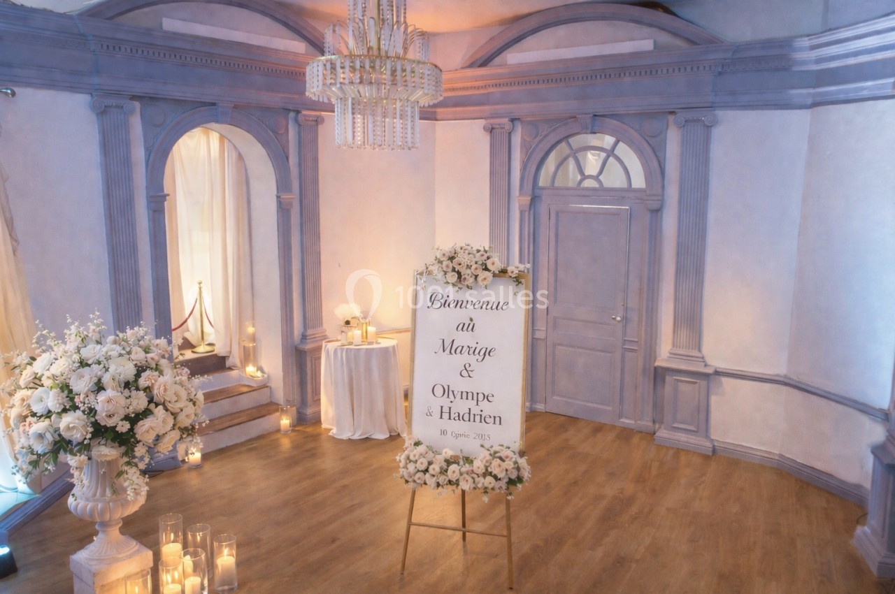 Salle décorée pour un mariage avec un panneau de bienvenue entouré de fleurs et des bougies disposées au sol.