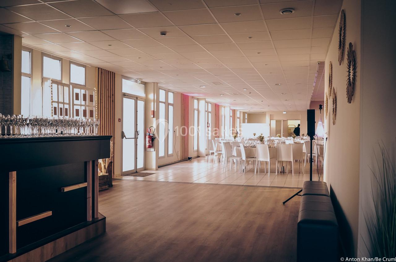 Salle de réception lumineuse avec tables dressées, chaises blanches et un bar à gauche.