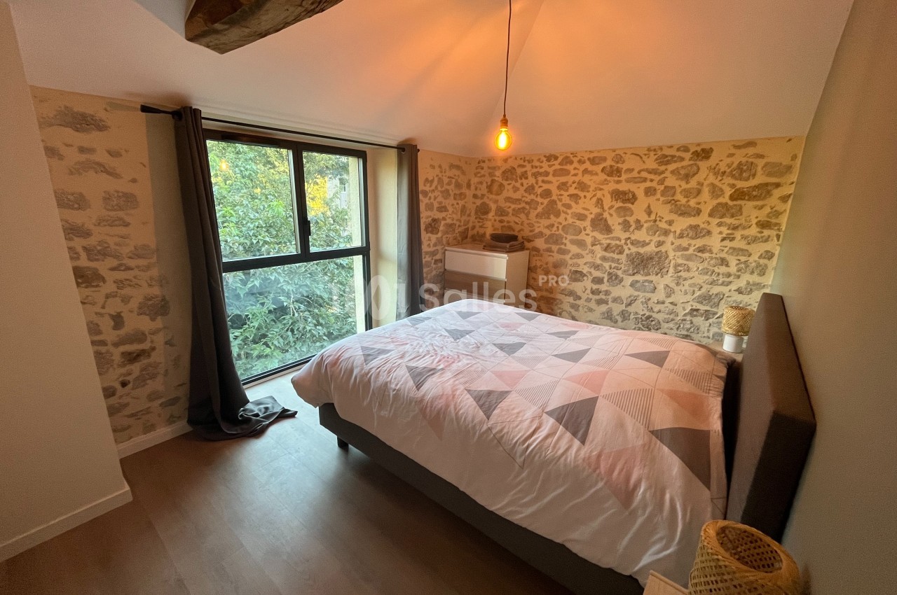 Chambre avec mur en pierre apparente, lit double, grande fenêtre donnant sur un jardin et éclairage suspendu.