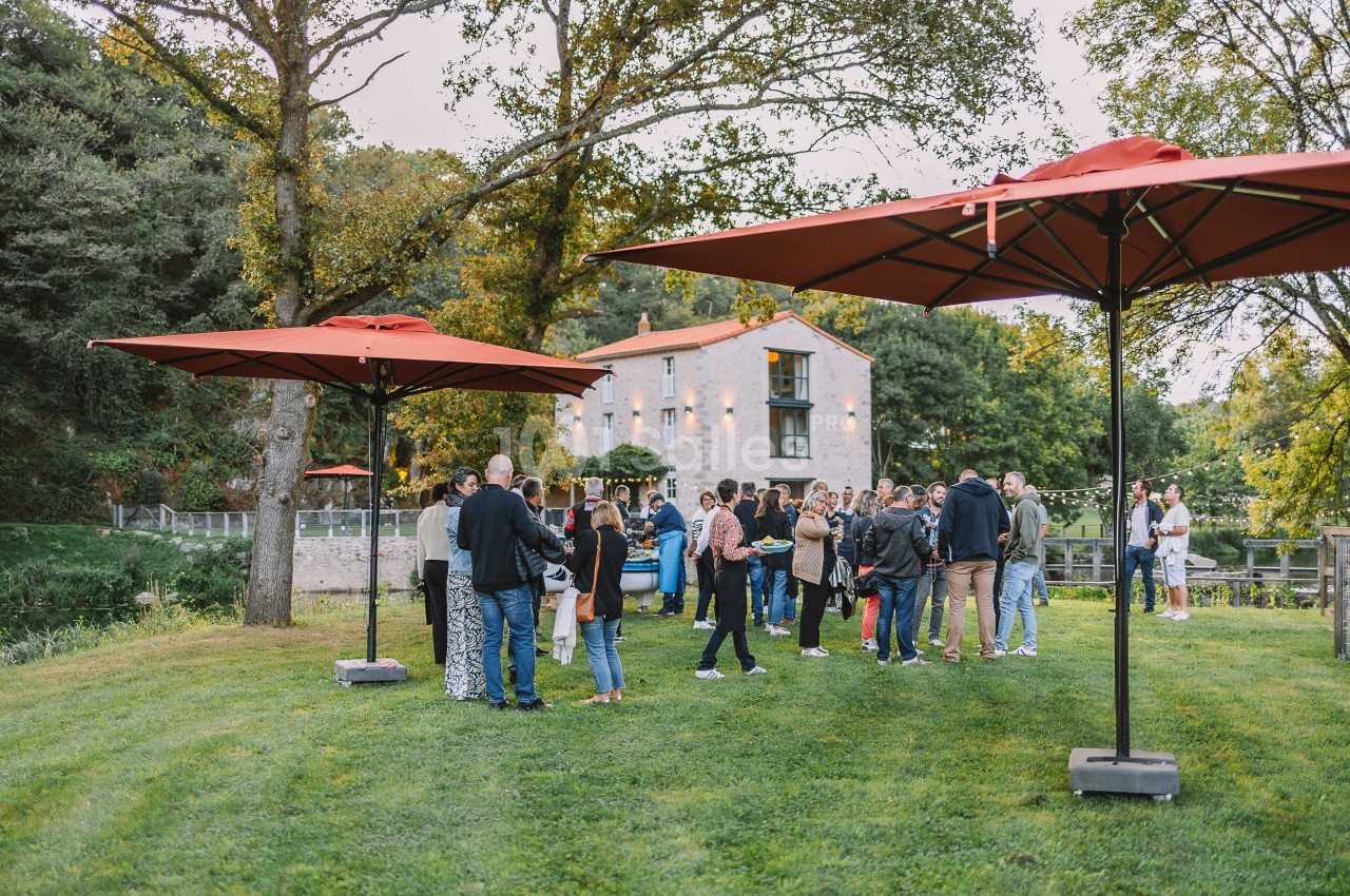 Groupe de personnes rassemblées sur une pelouse près d'un bâtiment en pierre, entouré d'arbres et de parasols rouges.