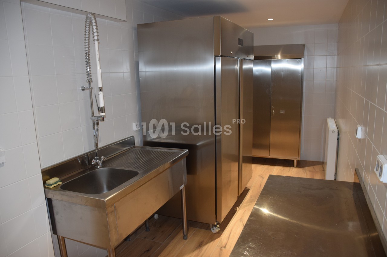 Cuisine professionnelle avec évier en inox, réfrigérateurs métalliques et murs carrelés blancs.