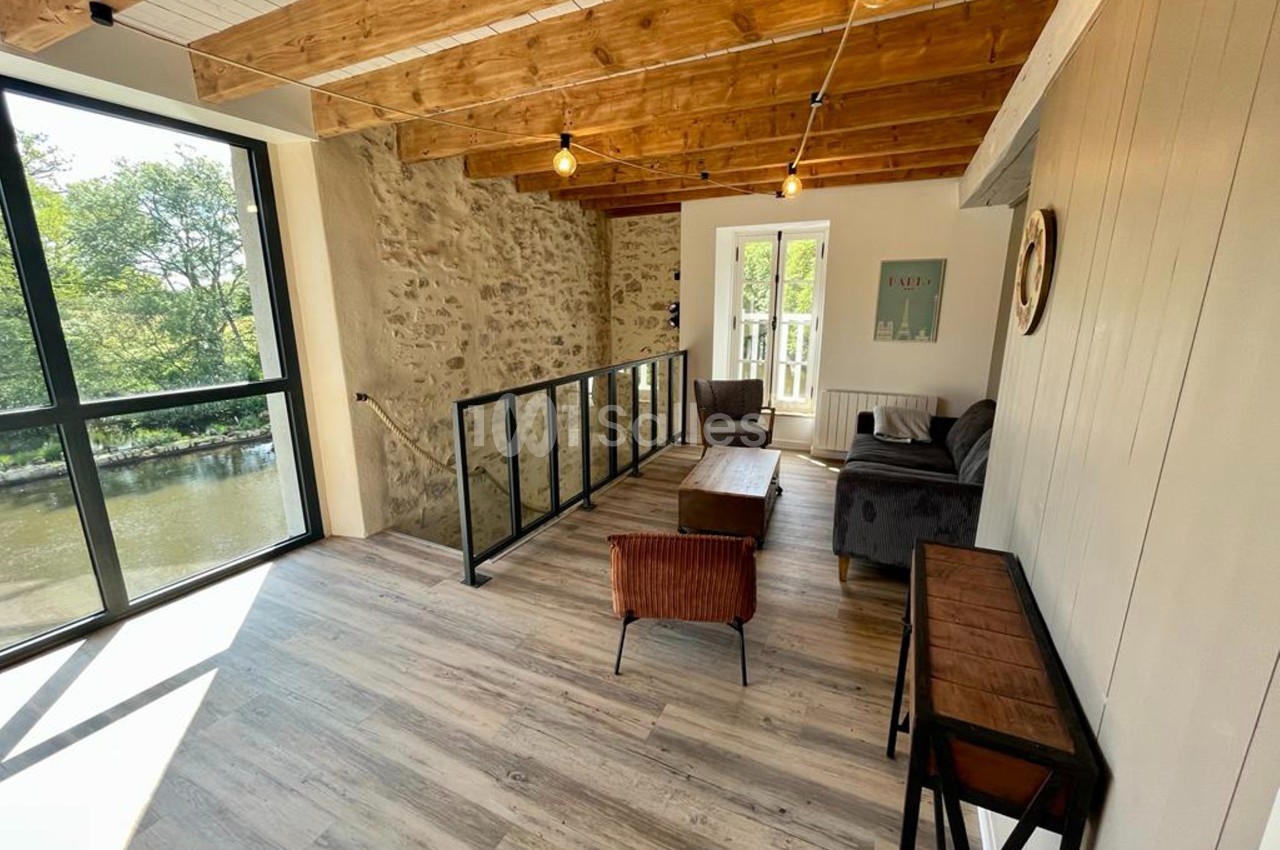Espace lumineux avec parquet, murs en pierre, poutres apparentes, baies vitrées et mobilier moderne, vue sur un cours d'eau.