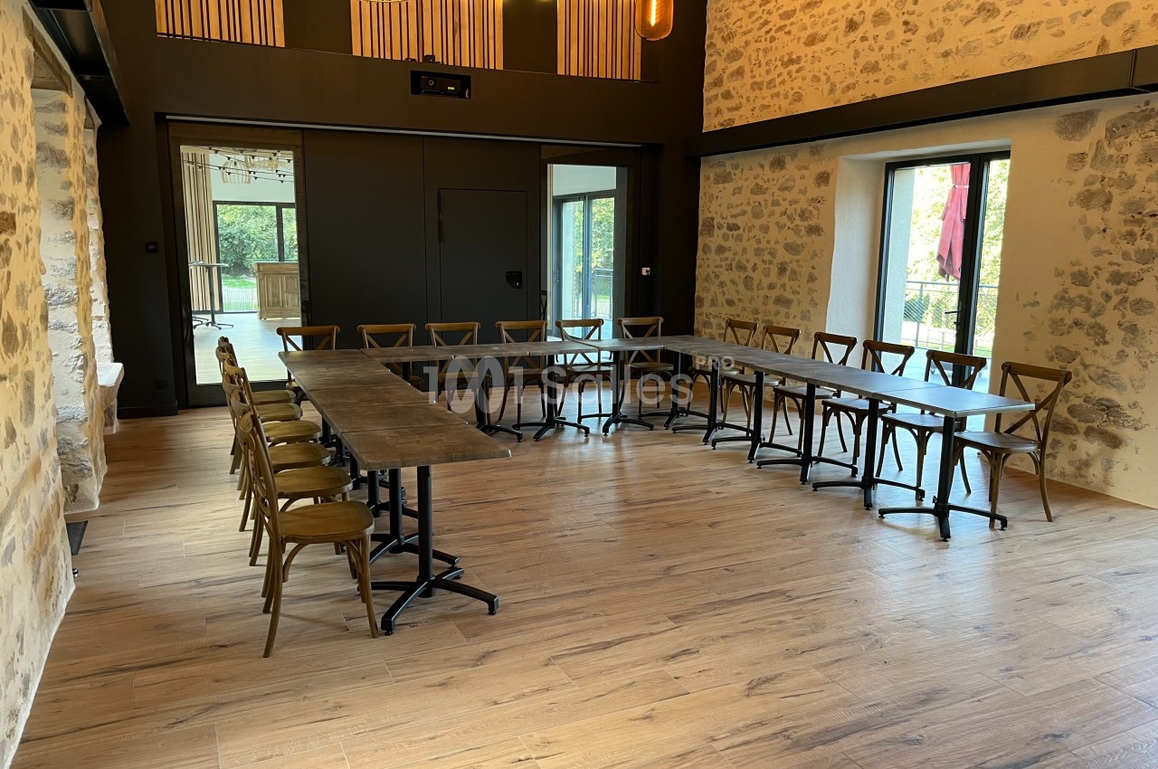 Salle de réunion lumineuse avec des murs en pierre, parquet en bois et tables disposées en U entourées de chaises.