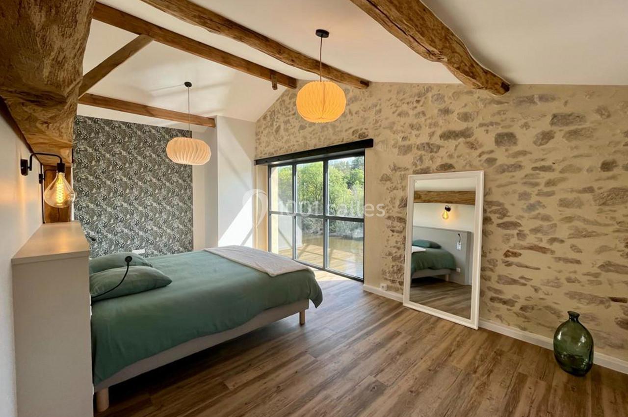 Chambre lumineuse avec lit double, murs en pierre apparente, parquet en bois et grande baie vitrée donnant sur l'extérieur.
