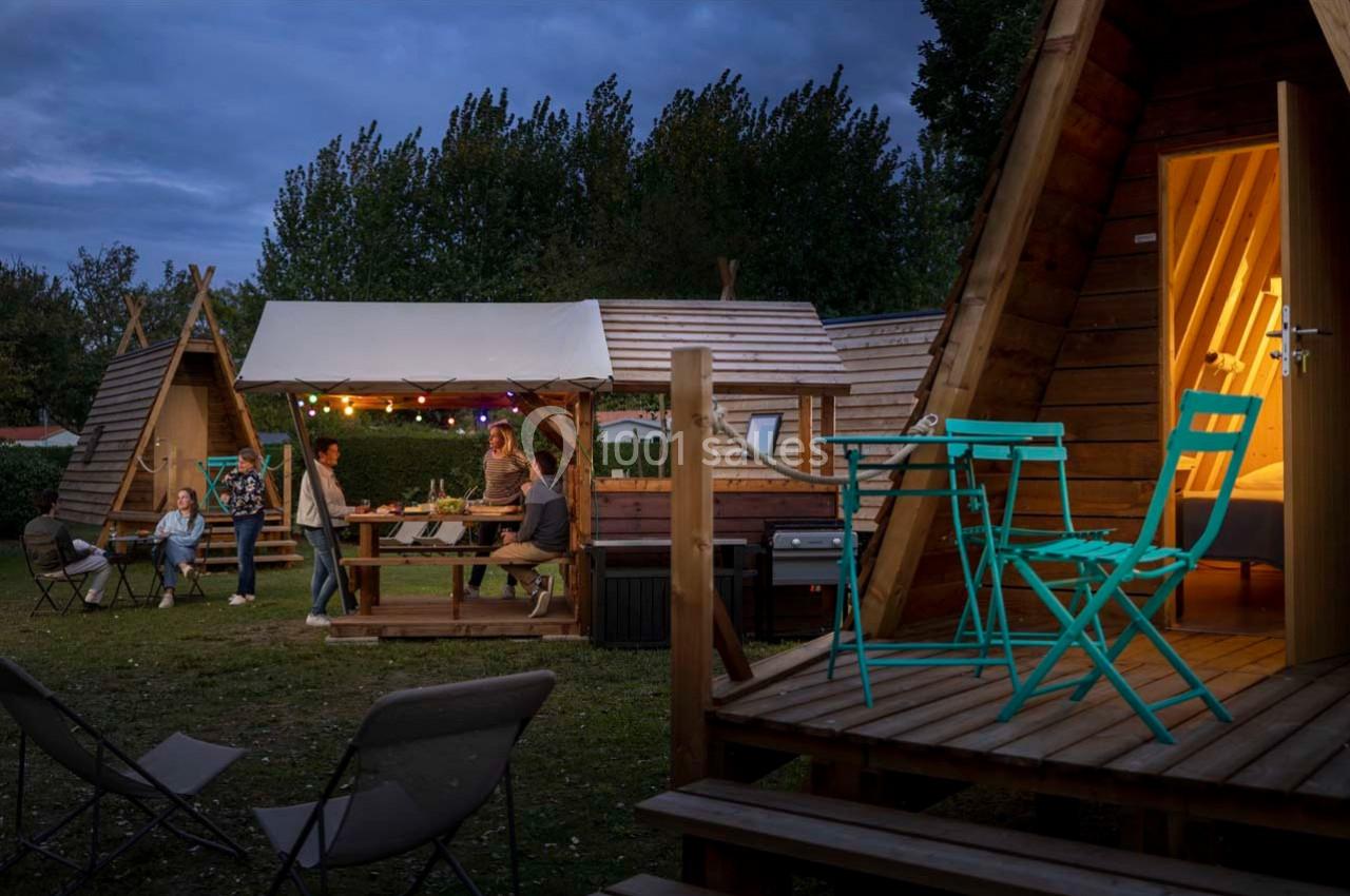 Des personnes dînent sous un auvent éclairé près de cabanes en bois triangulaires, entourées de mobilier de jardin.