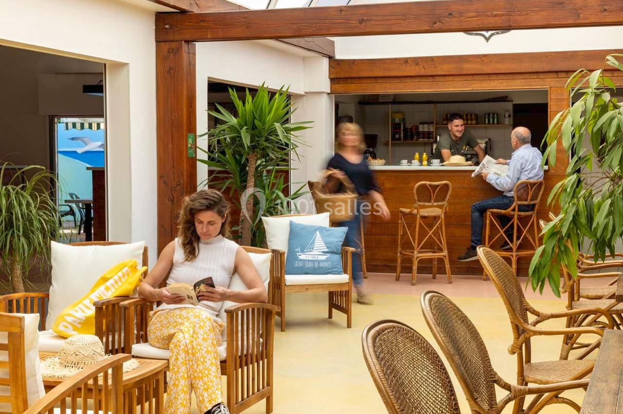 Un espace lumineux avec des plantes, des fauteuils en rotin, une femme lisant et un bar en arrière-plan.