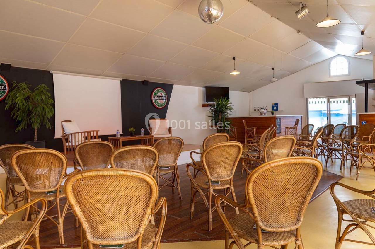 Salle aménagée avec des chaises en rotin, une petite scène, un bar en bois et des plantes décoratives.