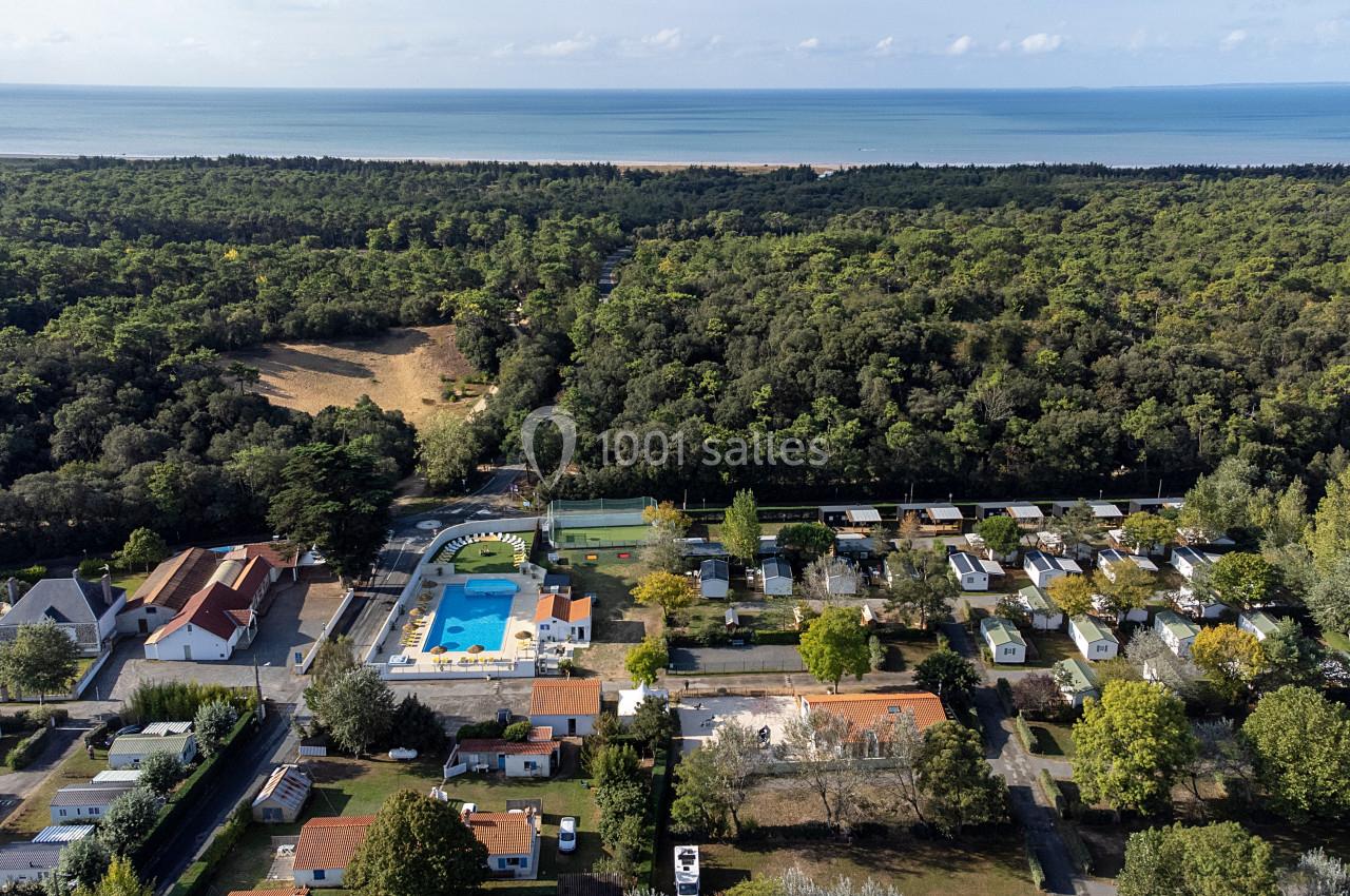Vue aérienne d'un camping avec piscine, mobil-homes et arbres, situé près d'une forêt et d'une plage en arrière-plan.