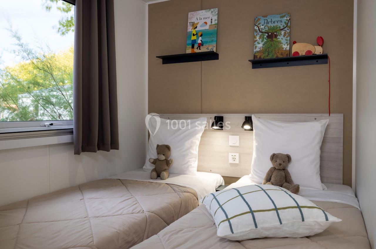 Deux lits simples avec des peluches, une fenêtre à gauche, et des étagères avec des livres au-dessus de la tête de lit.