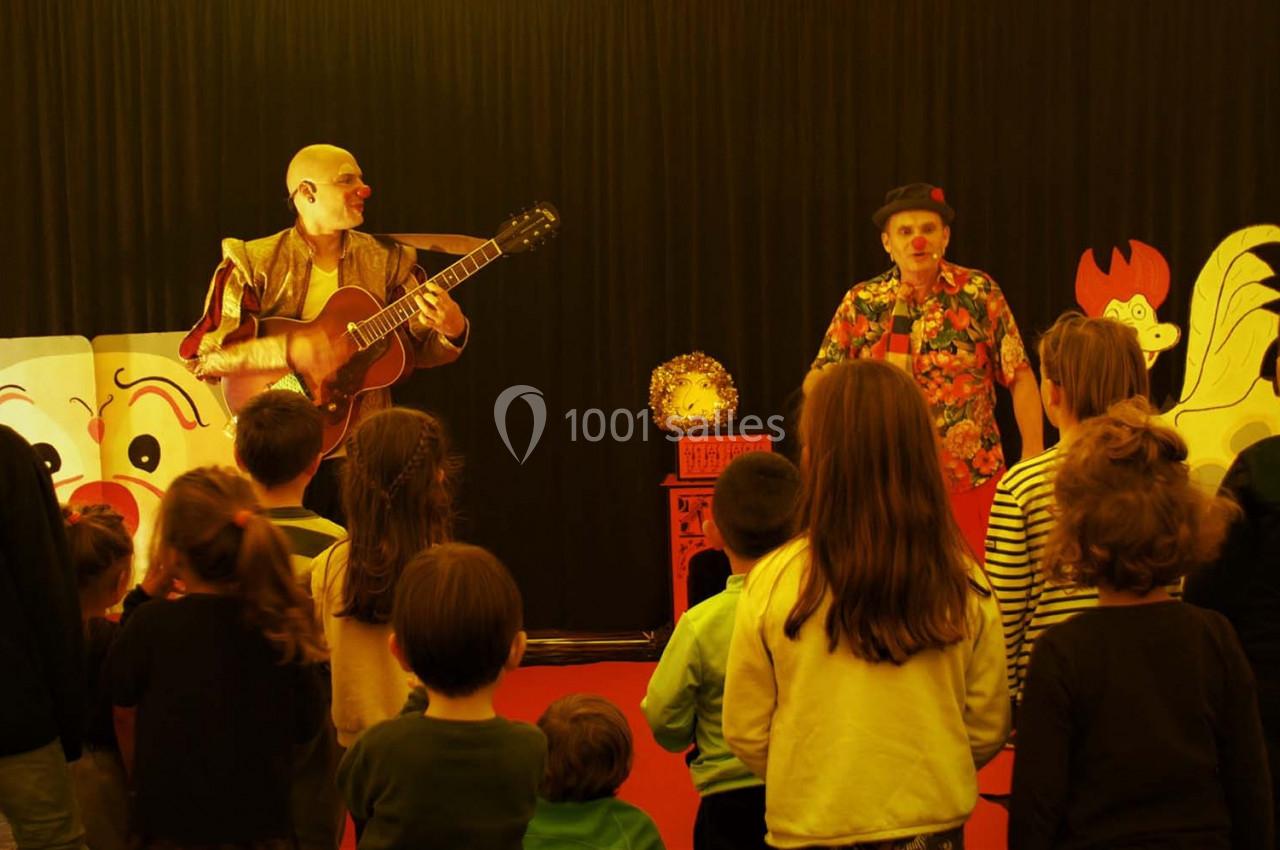 Des enfants assistent à un spectacle avec deux artistes, l’un jouant de la guitare, dans une salle décorée de motifs colorés.