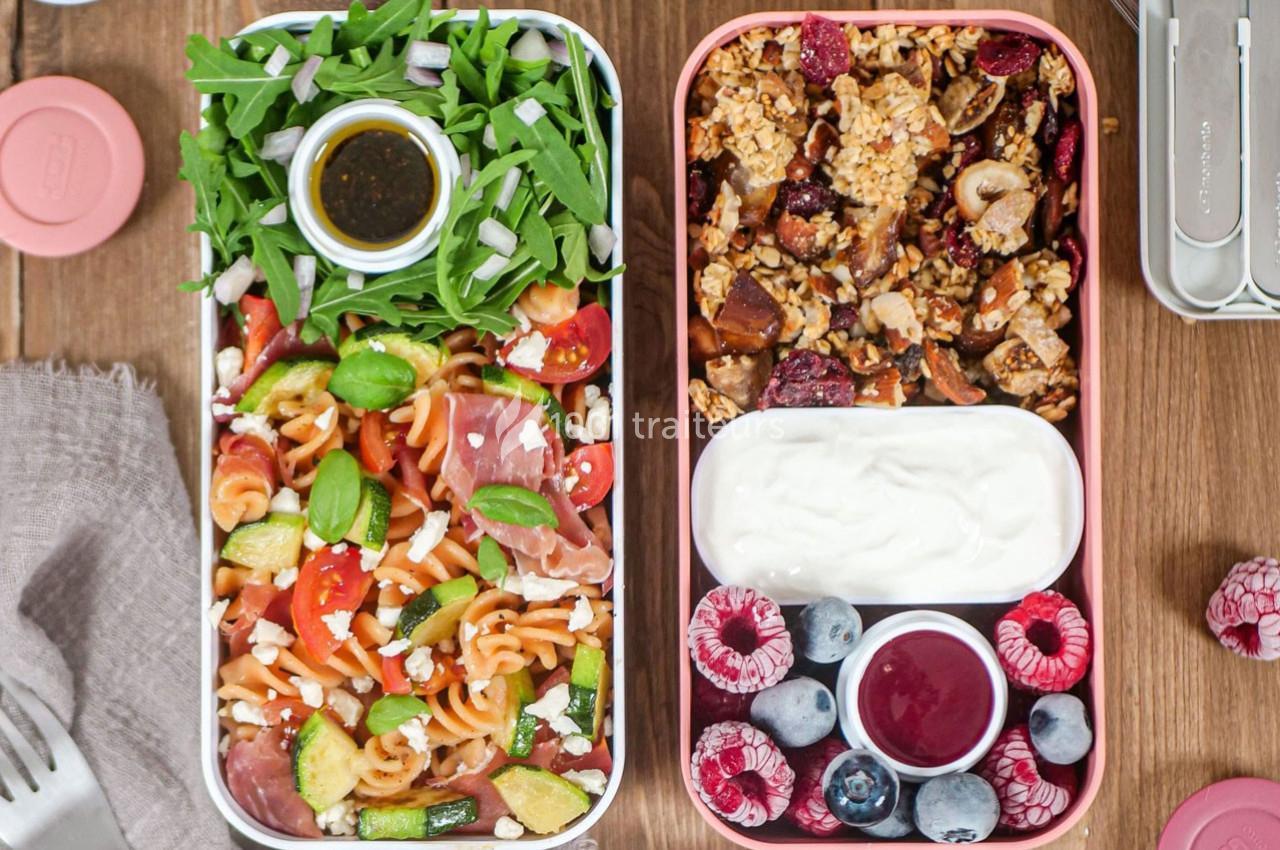Deux boîtes repas contenant des pâtes aux légumes, salade, granola, yaourt, fruits rouges et une sauce.