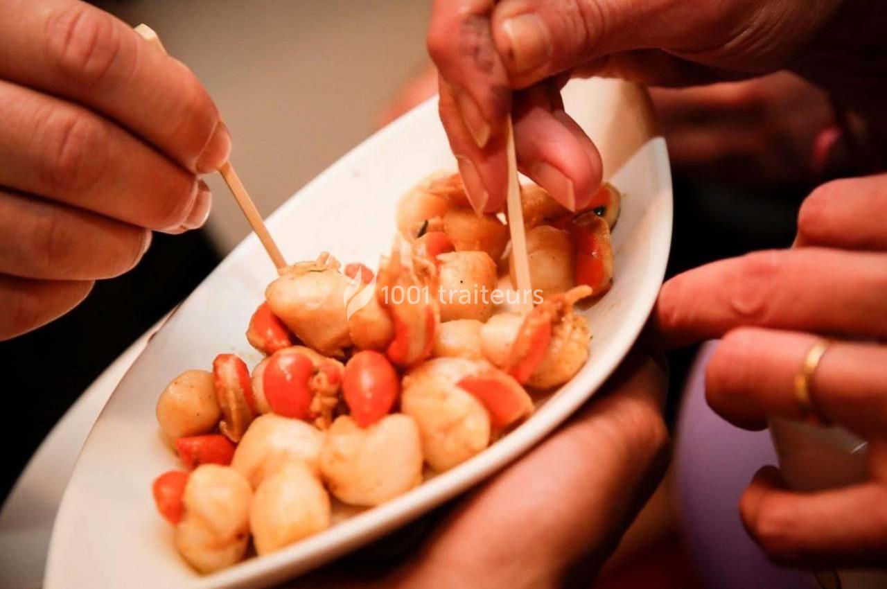 Des mains tiennent une assiette de brochettes apéritives composées de crevettes et tomates cerises.