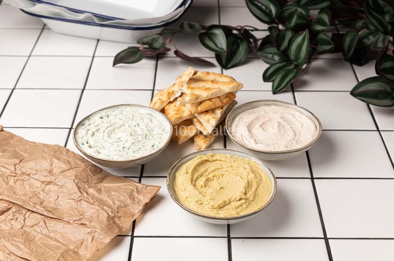 Trois bols de sauces (houmous, crème et herbes) avec des morceaux de pain plat sur une table carrelée blanche.