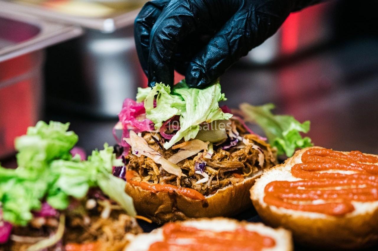 Un cuisinier garnit un burger avec de la salade et du chou sur une base de viande effilochée et de sauce.