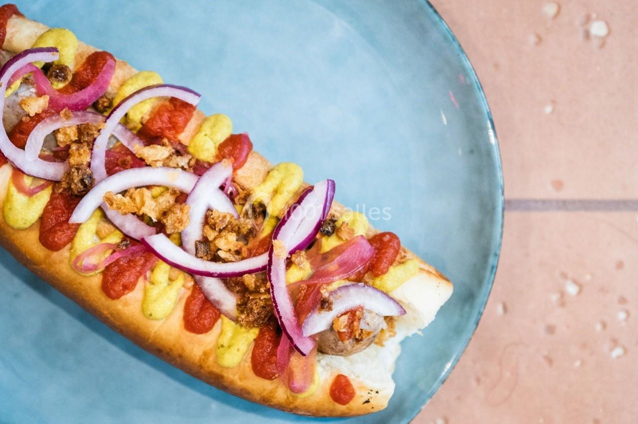 Hot-dog garni de moutarde, ketchup, oignons rouges et oignons frits, présenté sur une assiette bleue.